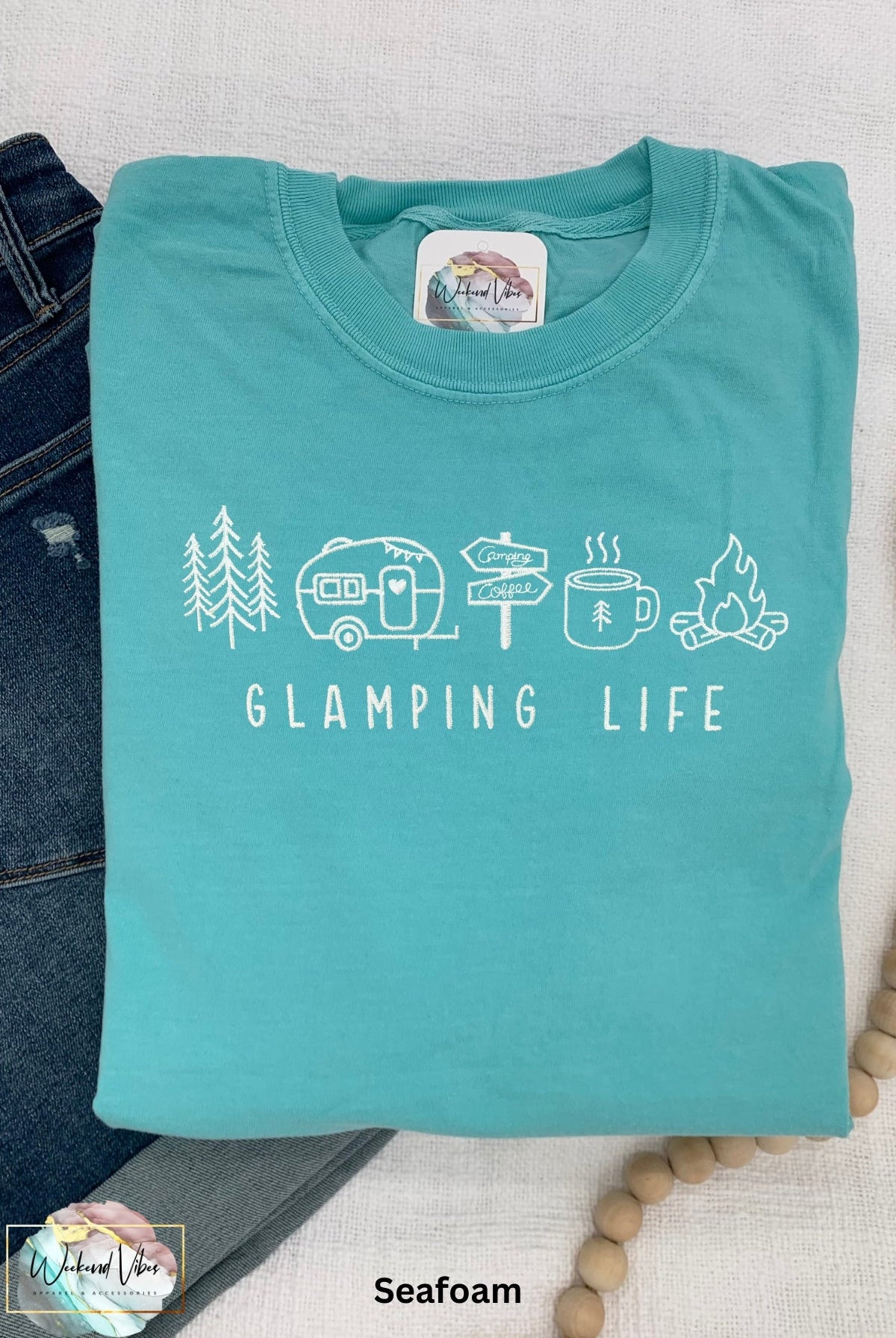 A embroidered t-shirt in seafoam color with 'GLAMPING LIFE' text and small illustrations of a camping scene, including trees, a ravel trailer, signs, coffee cup, and campfire.