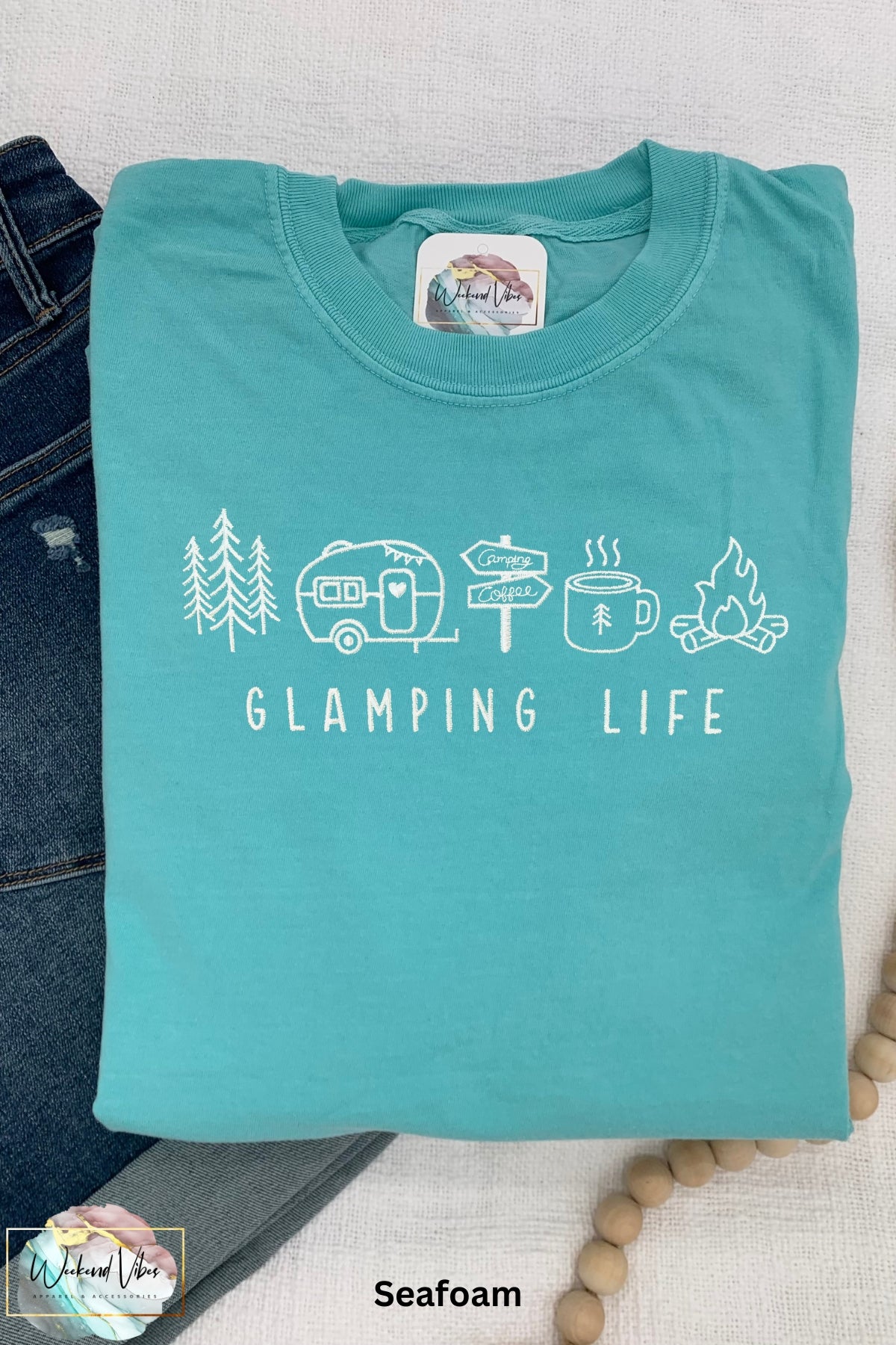 A embroidered t-shirt in seafoam color with 'GLAMPING LIFE' text and small illustrations of a camping scene, including trees, a ravel trailer, signs, coffee cup, and campfire.