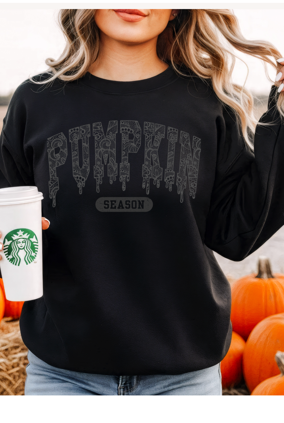 Person wearing a black sweatshirt with 'Pumpkin Season' text, holding a Starbucks cup, surrounded by pumpkins.