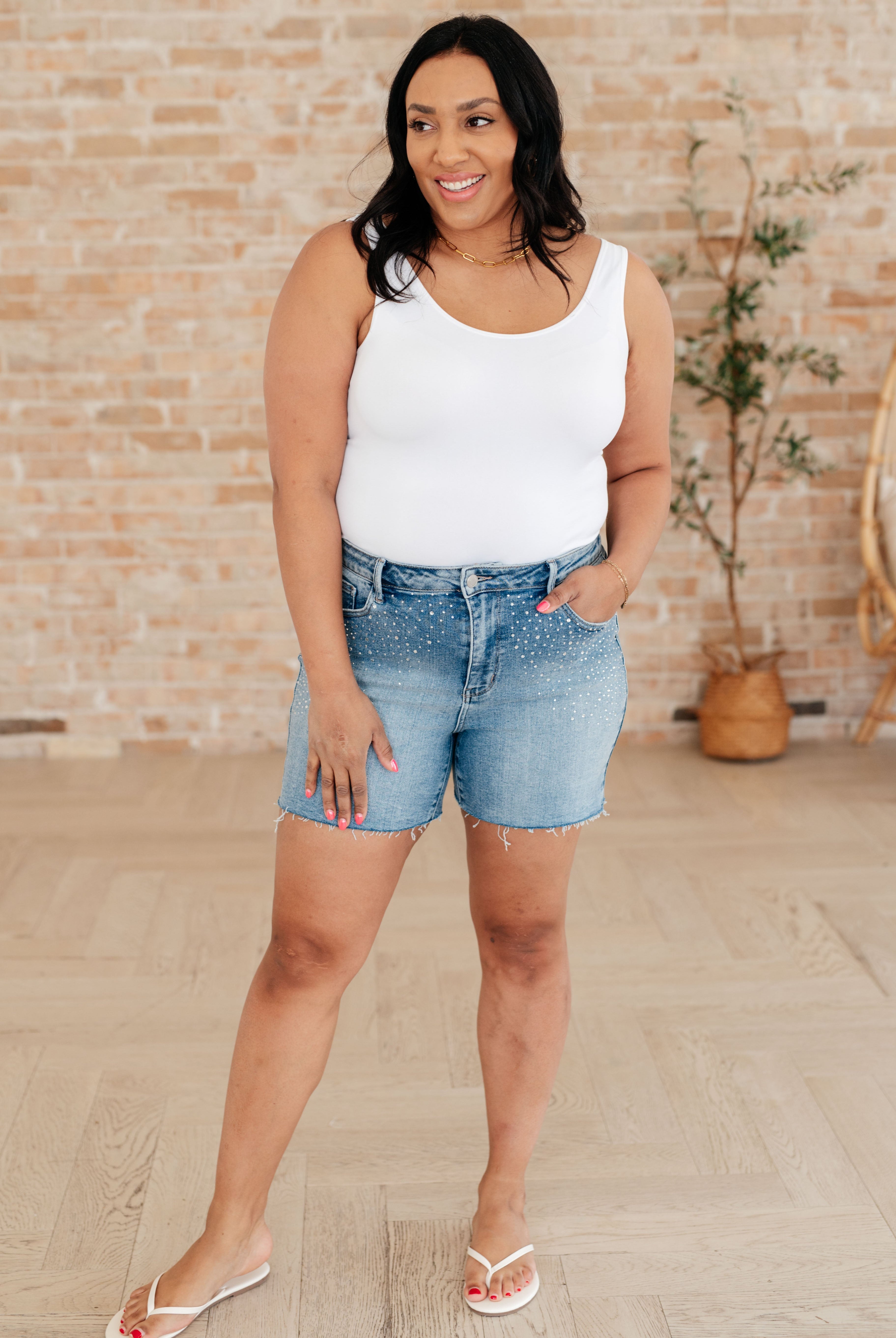 Krychele wearing a white tank top and denim shorts standing in front of a brick wall.