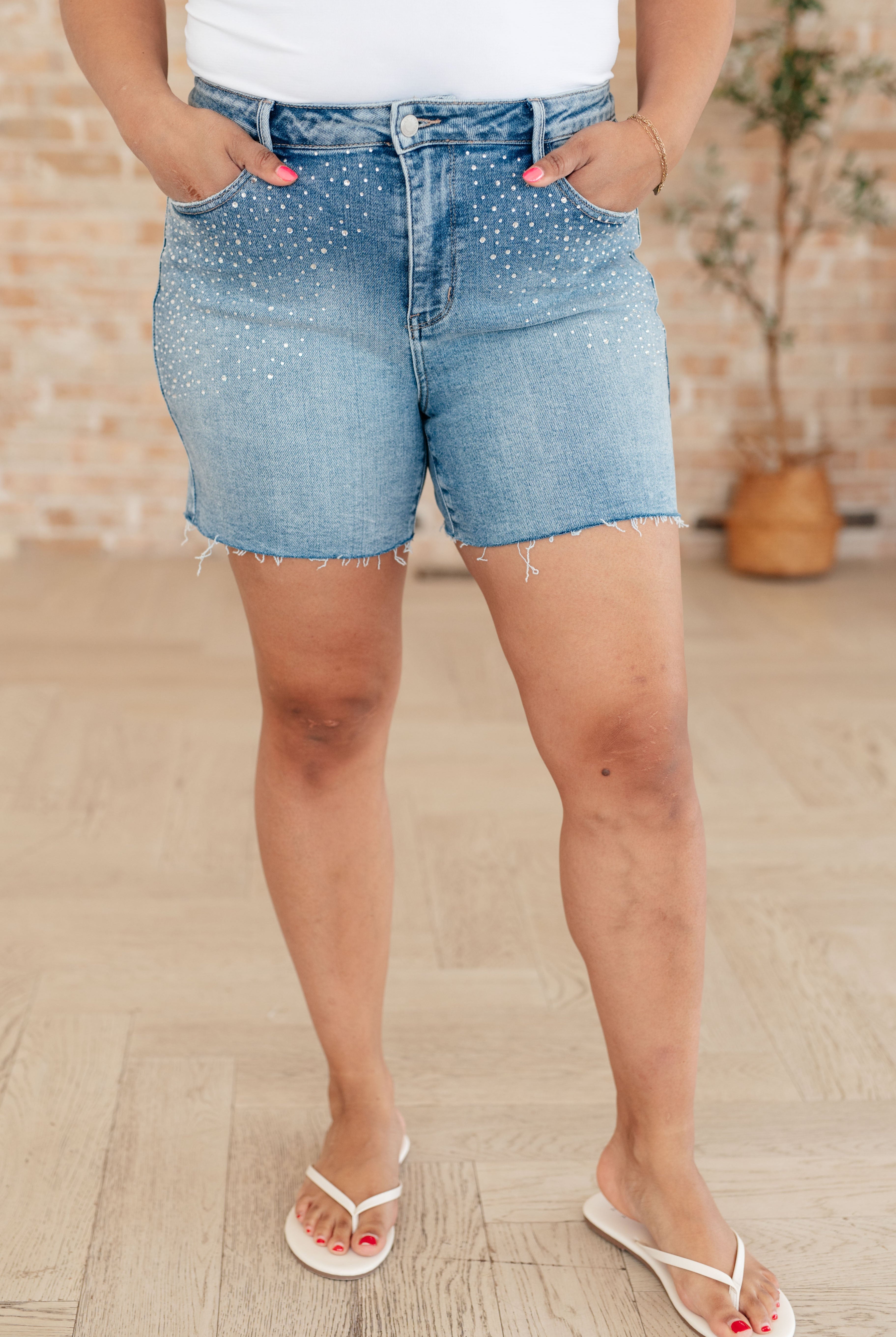 Model wearing raw edge cutoff Judy Blue blue denim shorts with rhinestones standing on wooden floor. Judy Blue Elle denim rhinestone shorts.