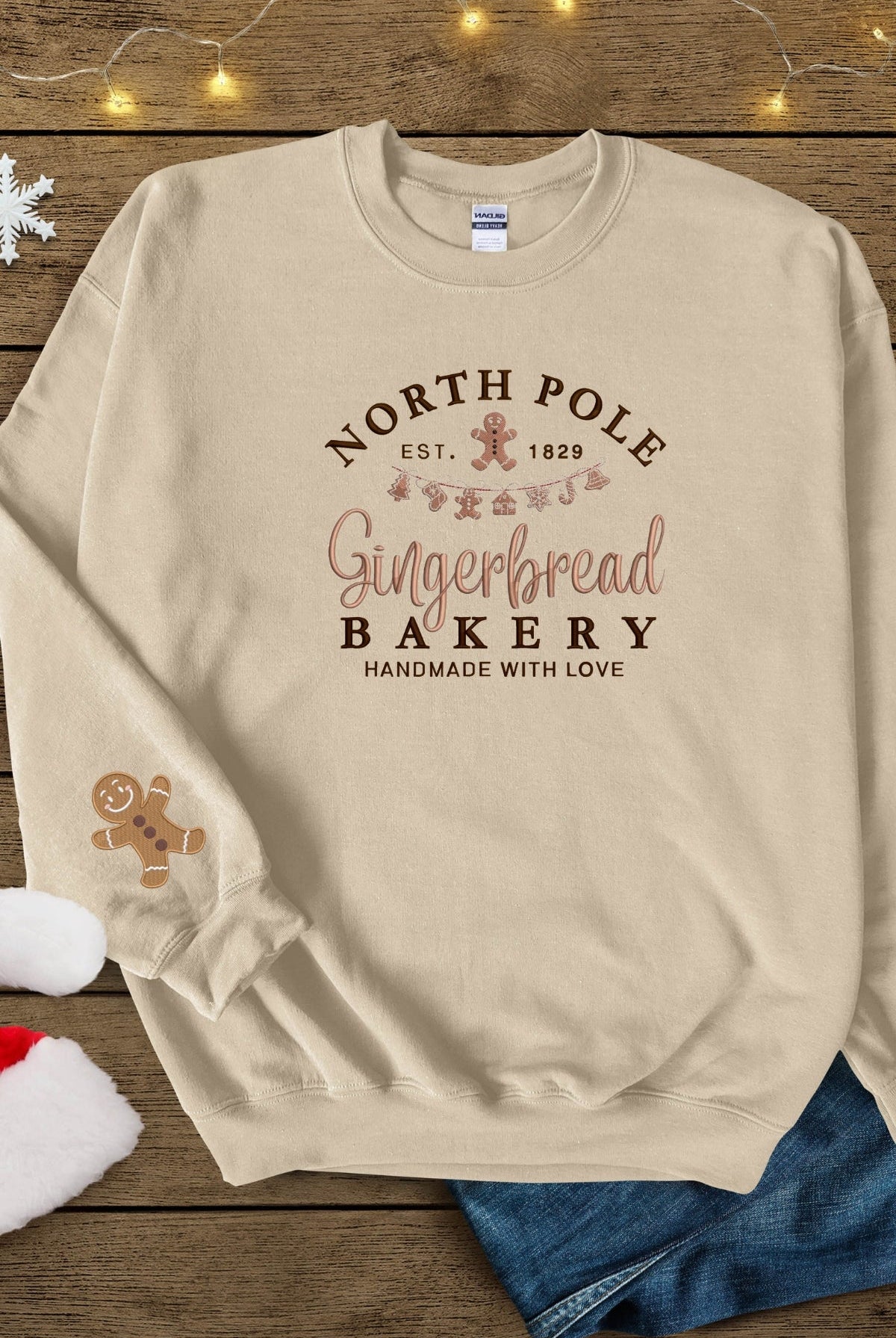 A sand colored beige sweatshirt with 'North Pole Gingerbread Bakery' embroidered on the chest and a small embroidered gingerbread man on the right sleeve.