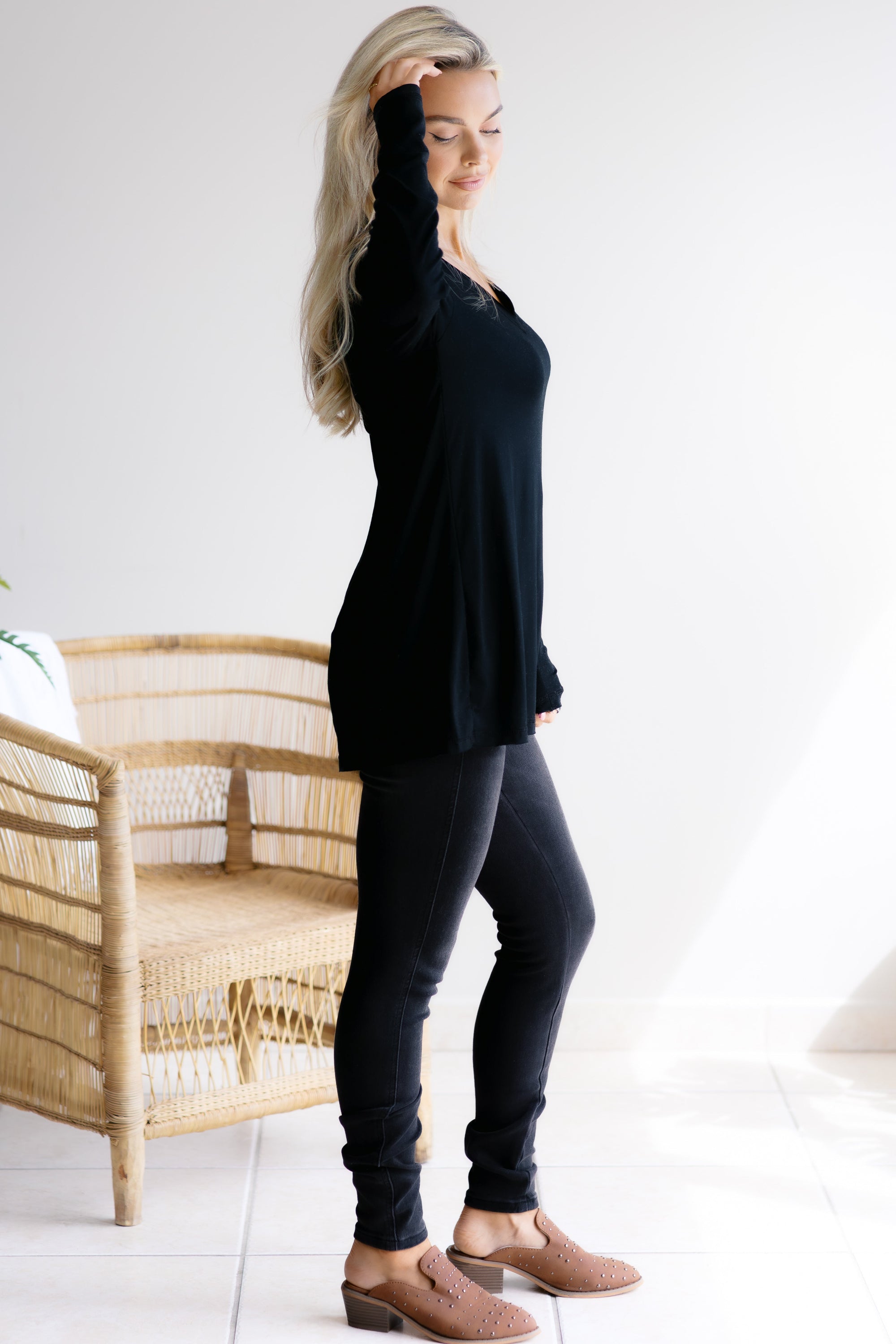 Woman wearing a black long-sleeve top and dark jeans standing in a room with a wicker chair.