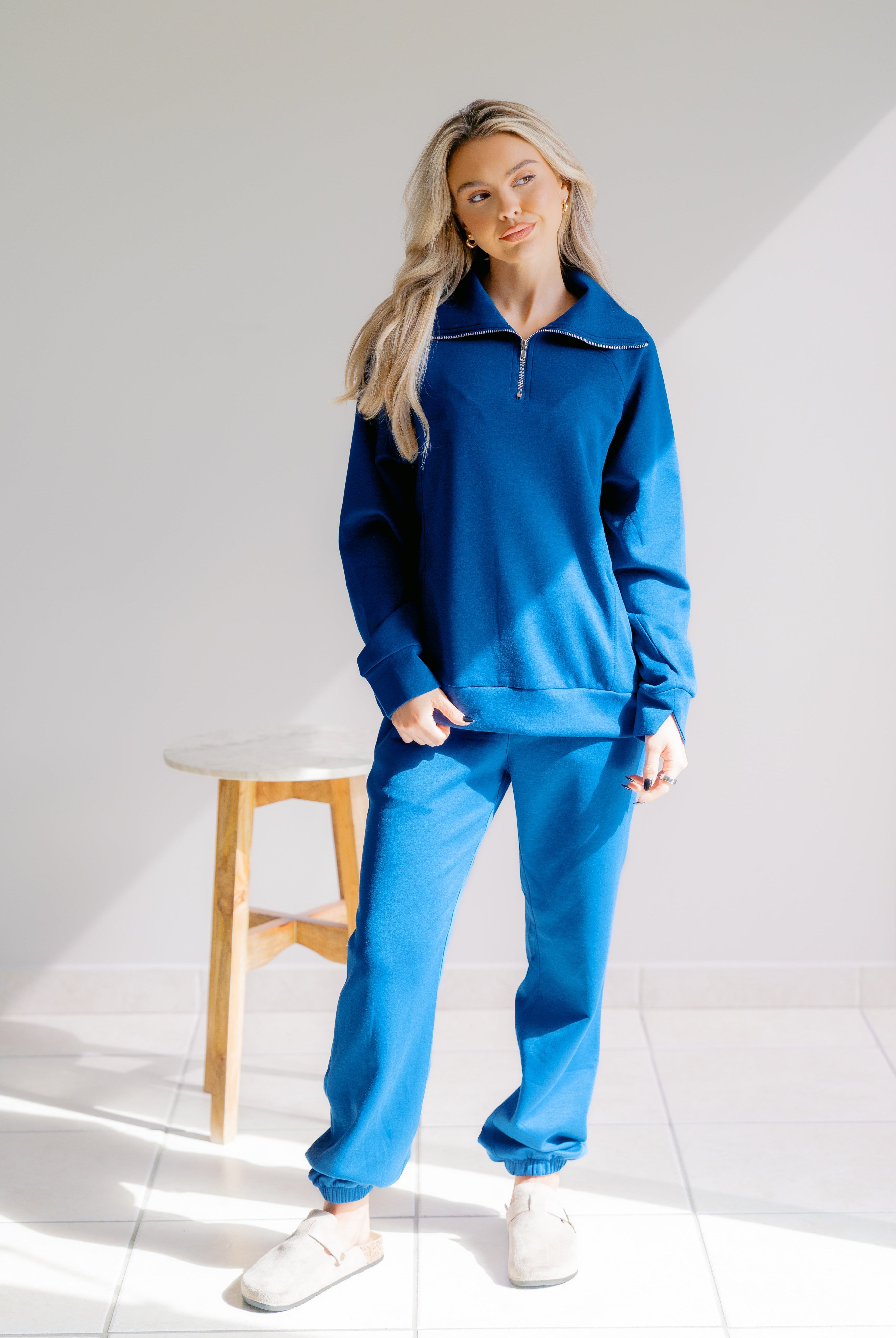 Woman wearing a blue tracksuit standing in a minimalistic room with a stool.