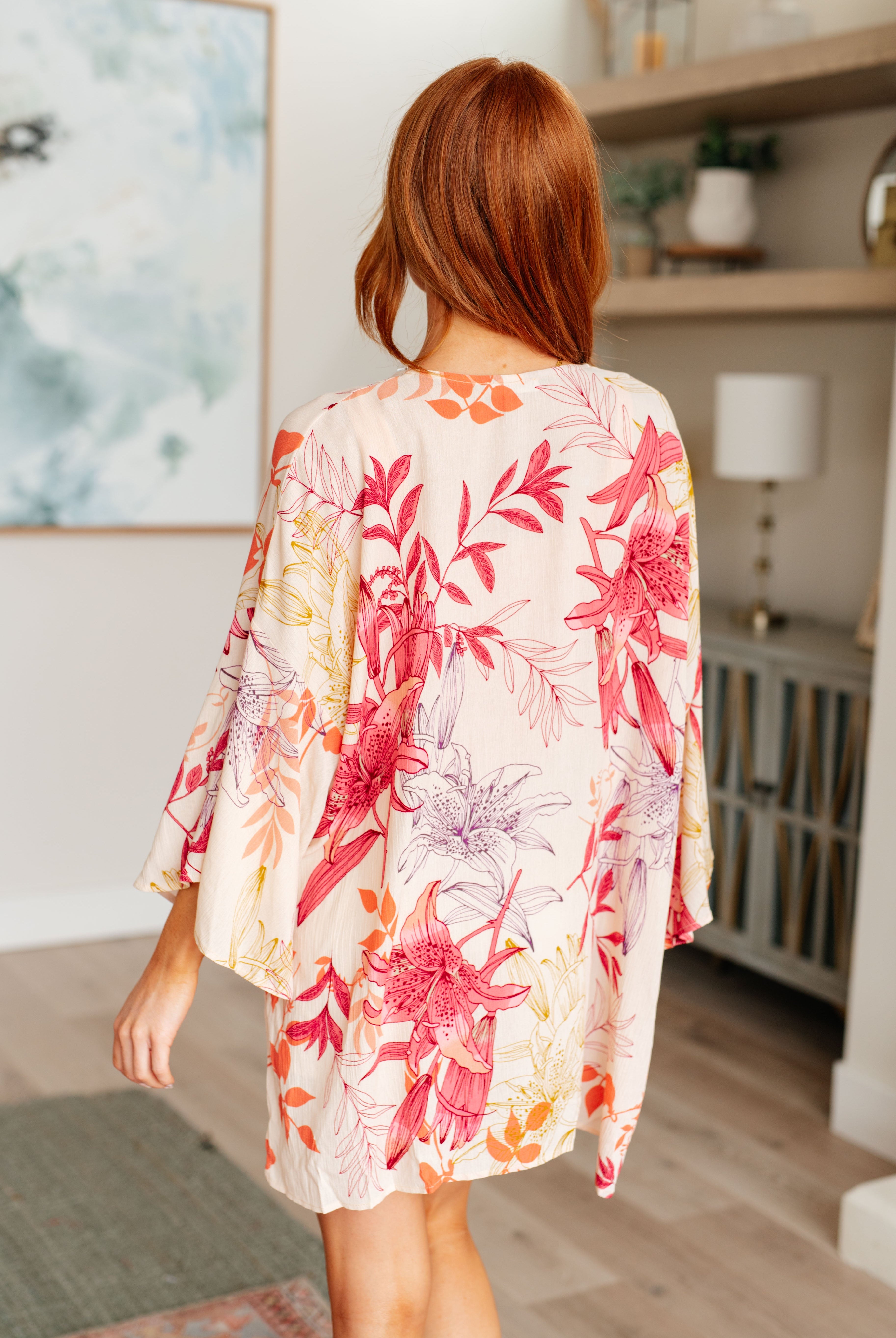 Woman wearing a floral kimono-style garment in a room with a painting and lamp in the background.