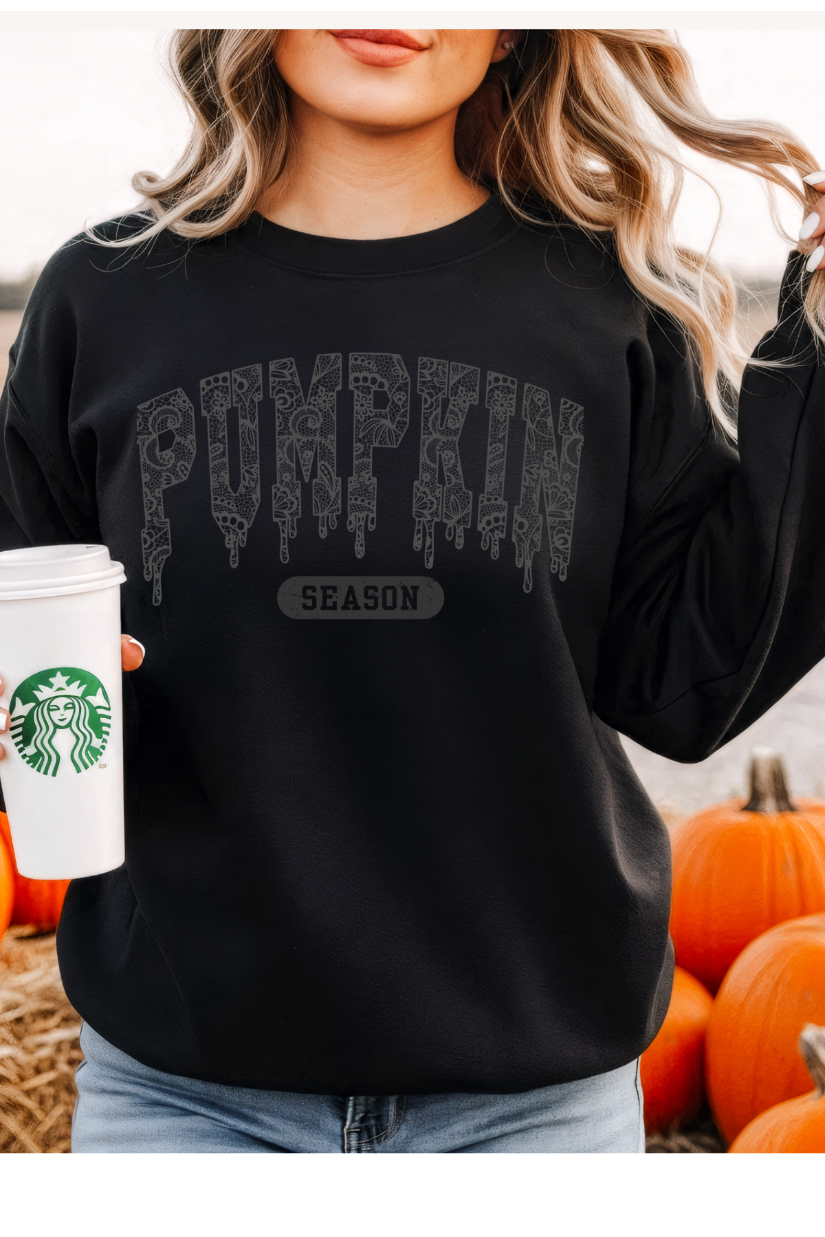 Person wearing a black sweatshirt with 'Pumpkin Season' text, holding a Starbucks cup, surrounded by pumpkins.