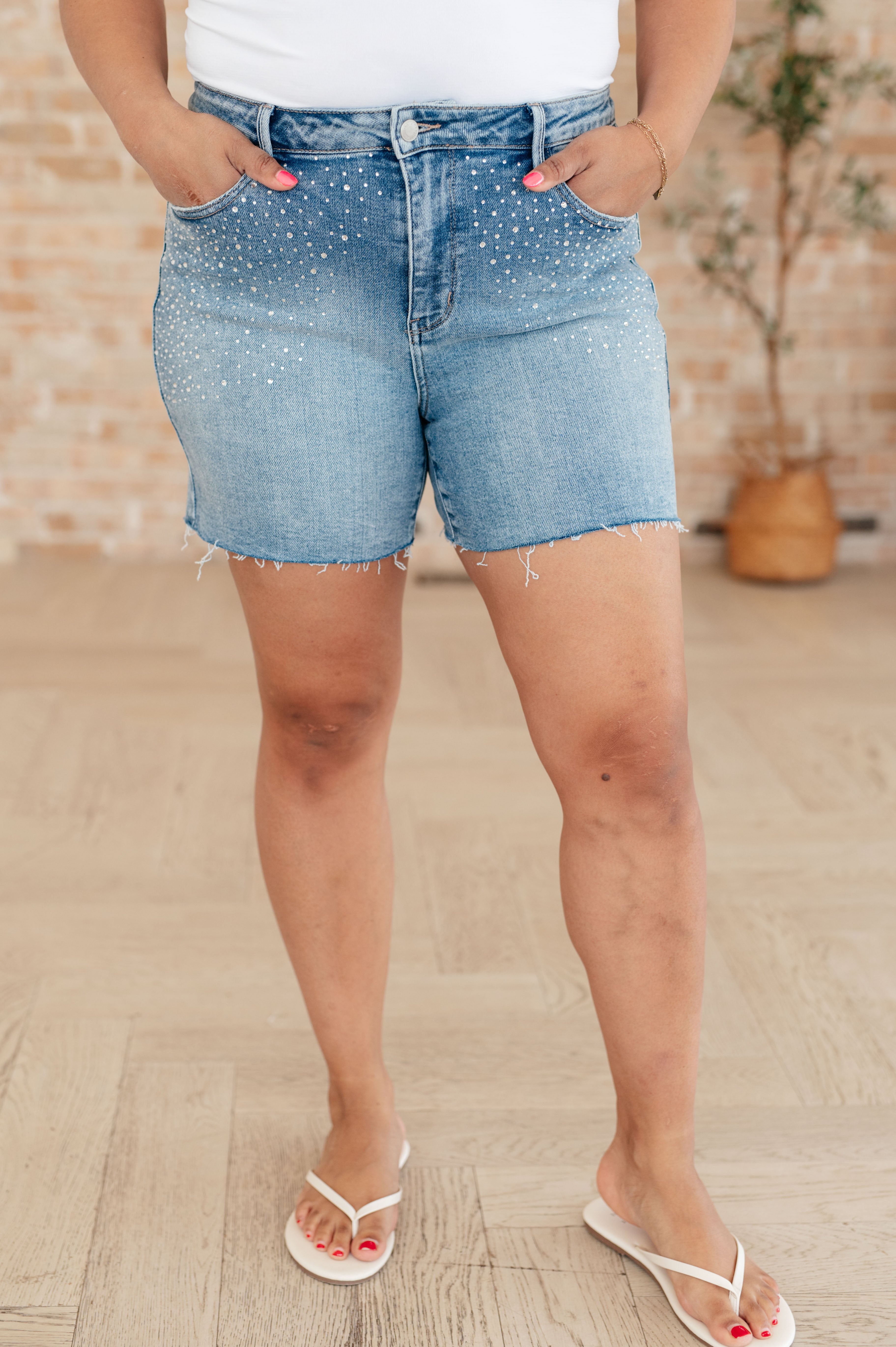 Model wearing raw edge cutoff Judy Blue blue denim shorts with rhinestones standing on wooden floor. Judy Blue Elle denim rhinestone shorts.