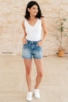 Woman wearing a white tank top and Judy Blue blue denim shorts standing against a brick wall.