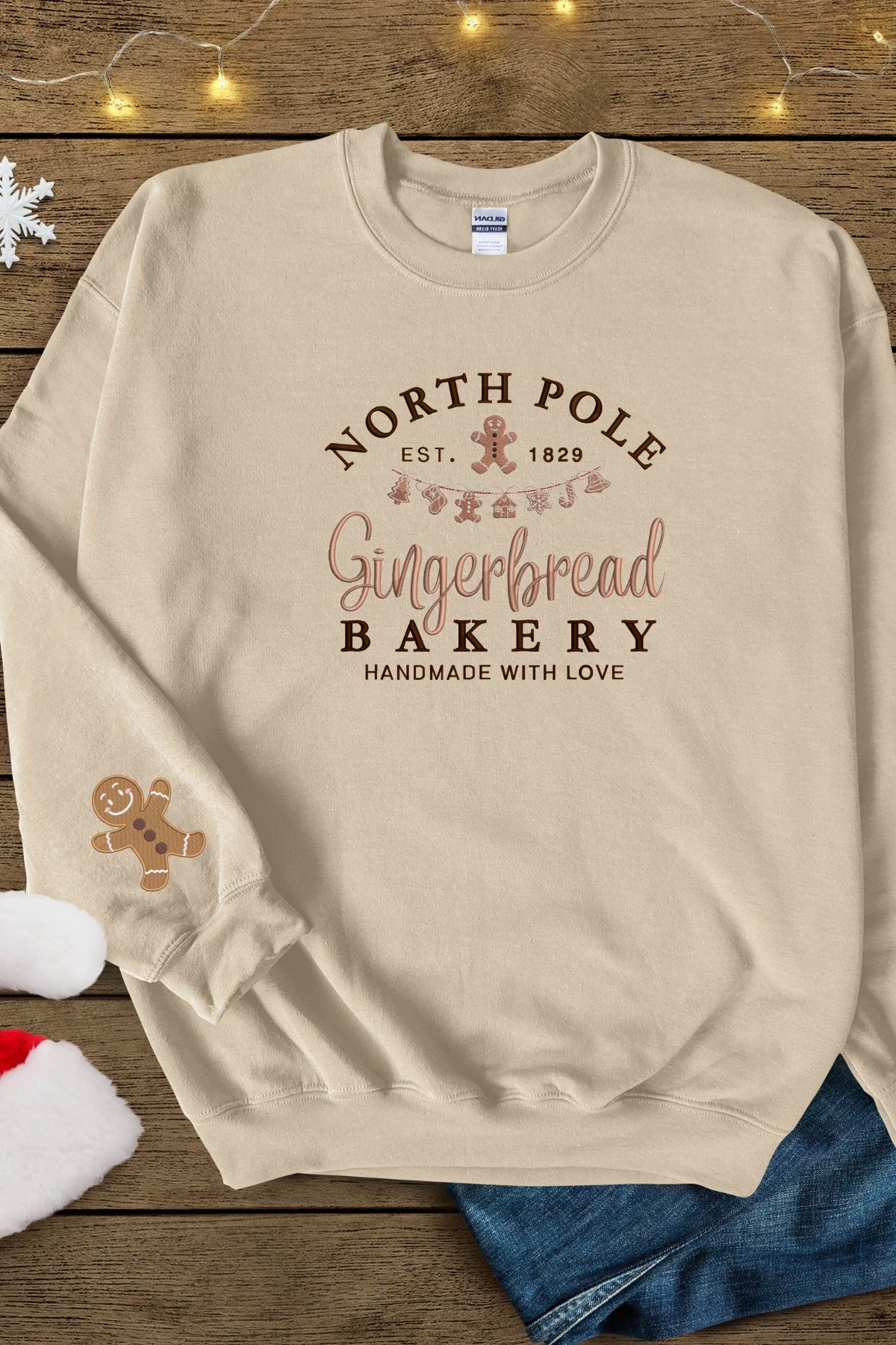 A sand colored beige sweatshirt with 'North Pole Gingerbread Bakery' embroidered on the chest and a small embroidered gingerbread man on the right sleeve.