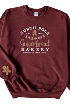 A cardinal red sweatshirt with 'North Pole Gingerbread Bakery' embroidered on the chest and a small embroidered gingerbread man on the right sleeve.