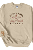 A beige sweatshirt with 'North Pole Gingerbread Bakery' embroidered on the chest and a small embroidered gingerbread man on the right sleeve.