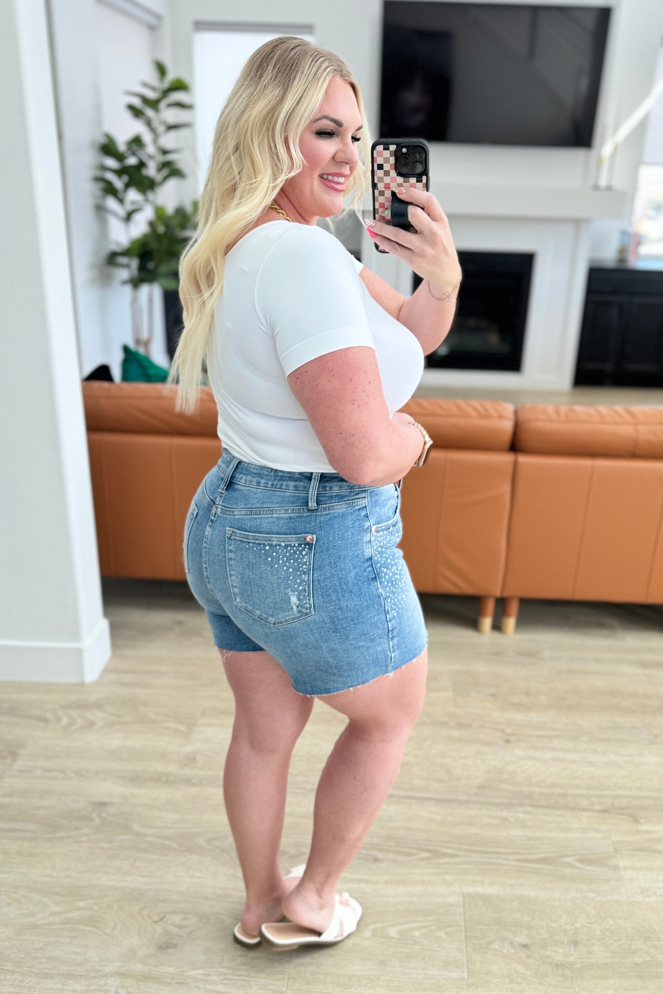 Kirra taking a mirror selfie wearing a white t-shirt and denim shorts in a living room.