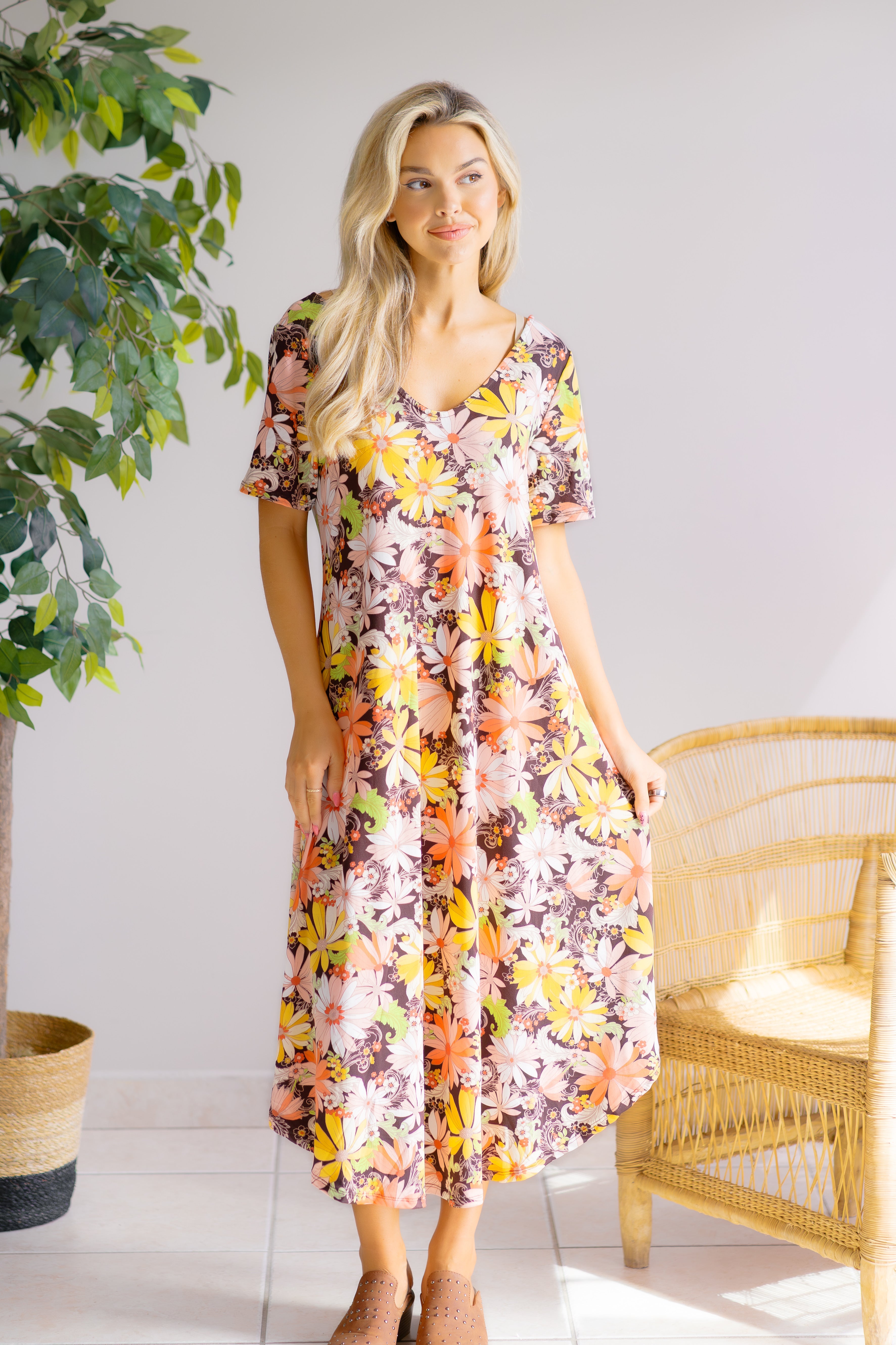 Woman wearing a fall colored, floral dress standing in a room with a plant and wicker chair.