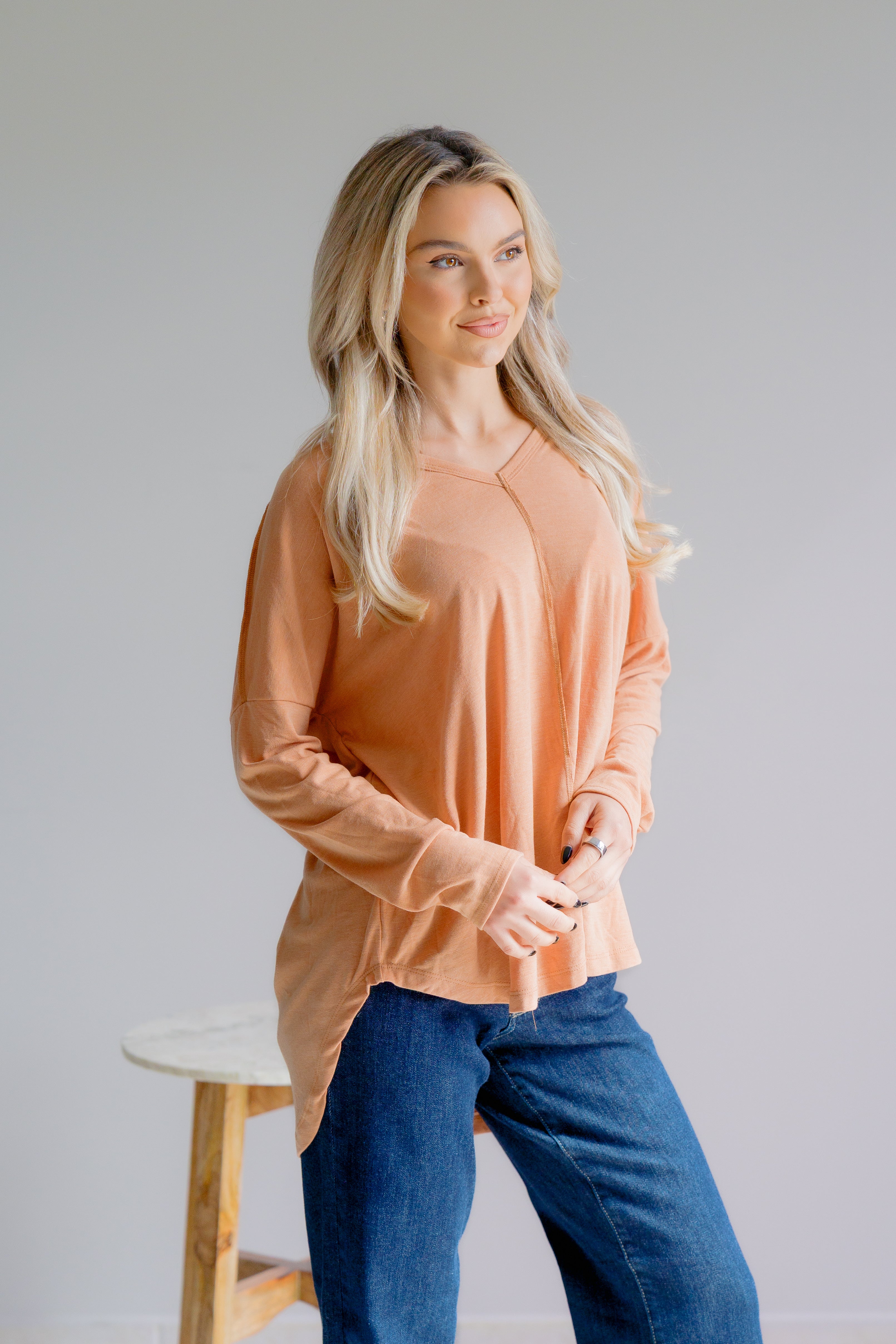 Woman wearing a peach long-sleeve top and blue jeans against a plain background