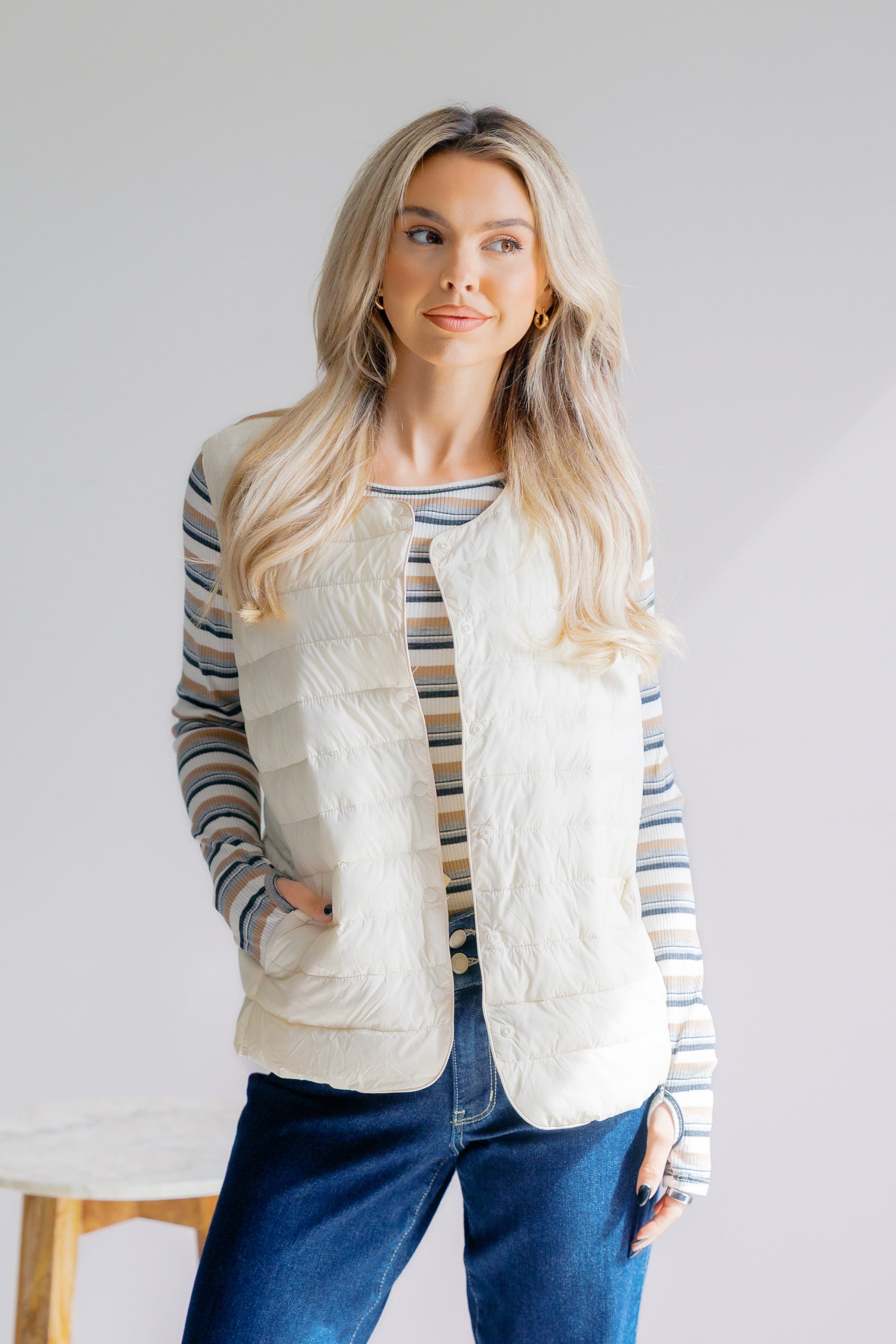 Woman wearing a striped shirt, white vest, and blue jeans against a plain background