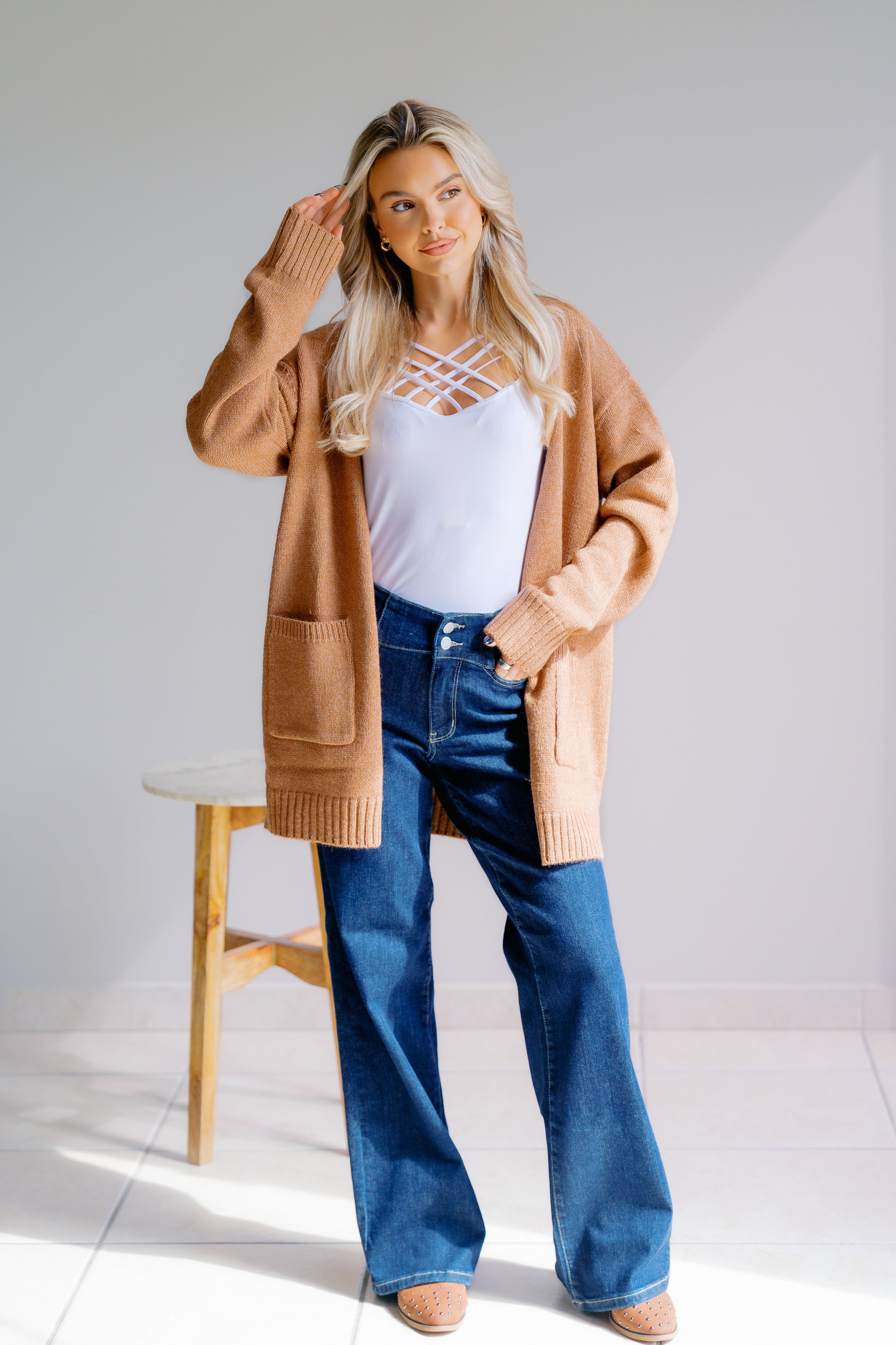 Woman wearing a beige cardigan paired with white criss cross tank and blue jeans in a minimalistic setting.