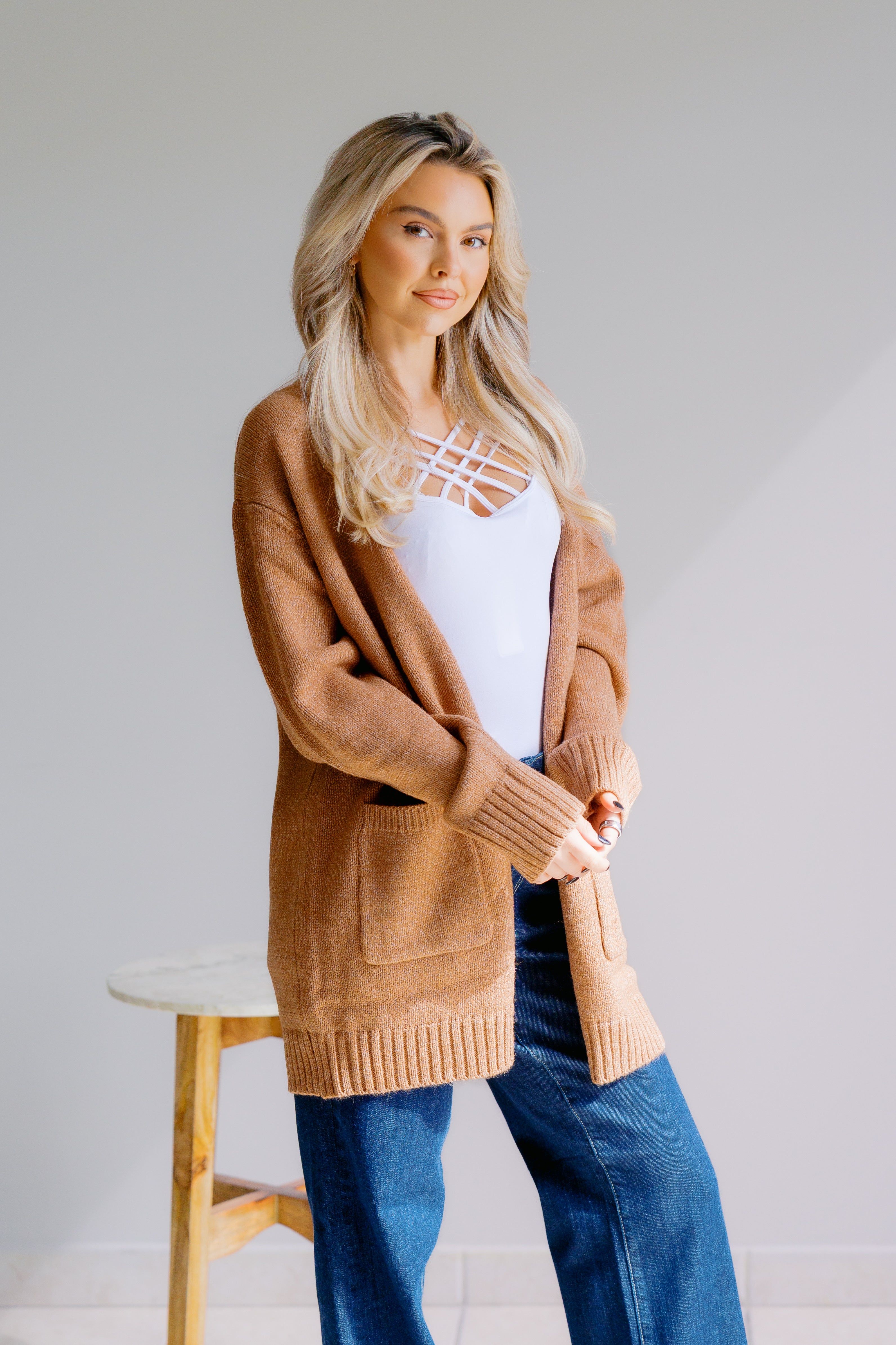 Woman wearing a brown cardigan over a white top and blue jeans on a plain background
