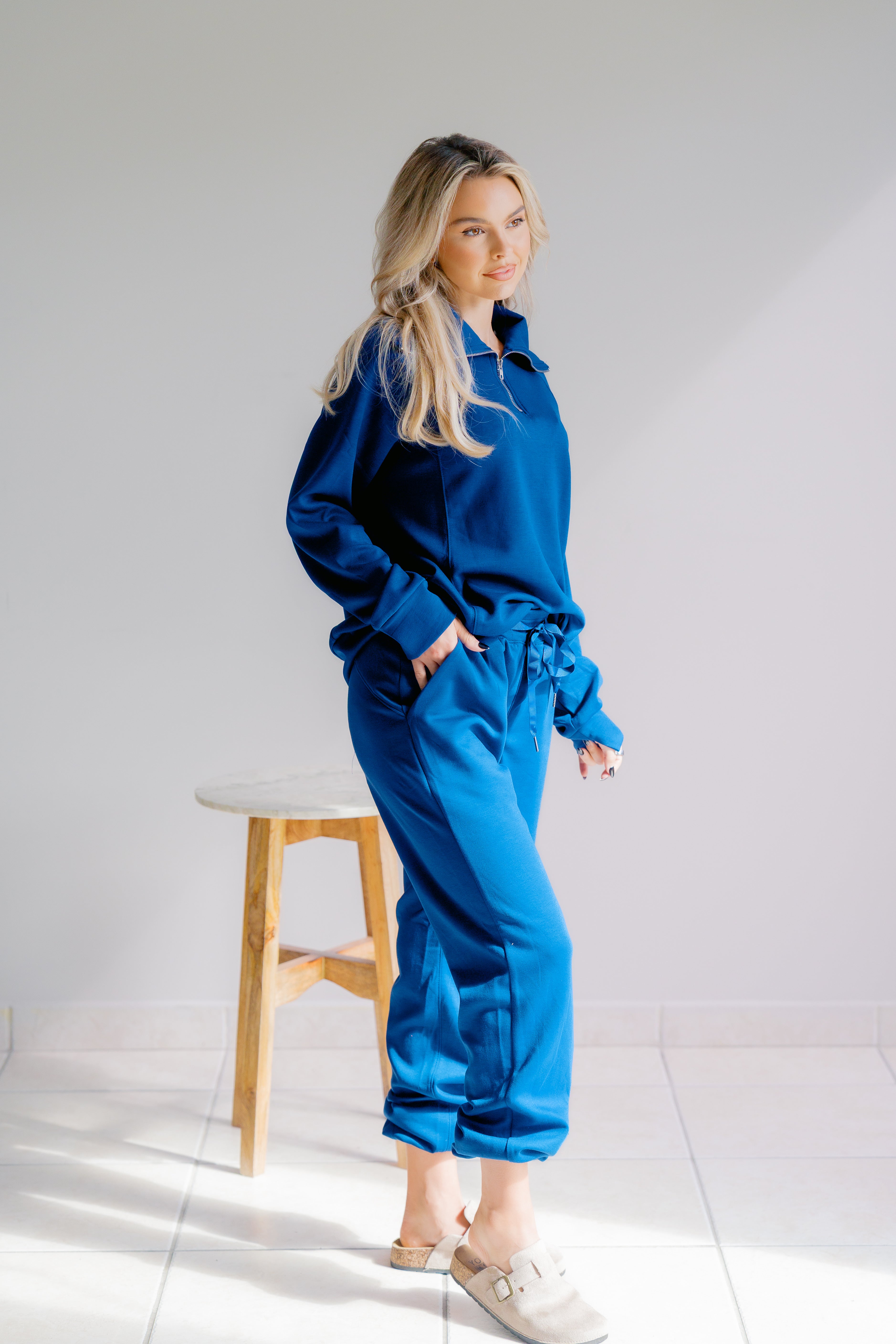 Woman in a blue tracksuit standing next to a wooden stool on a white background