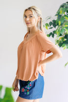 Woman wearing a peach blouse and blue skirt with floral design, standing against a white wall with greenery.