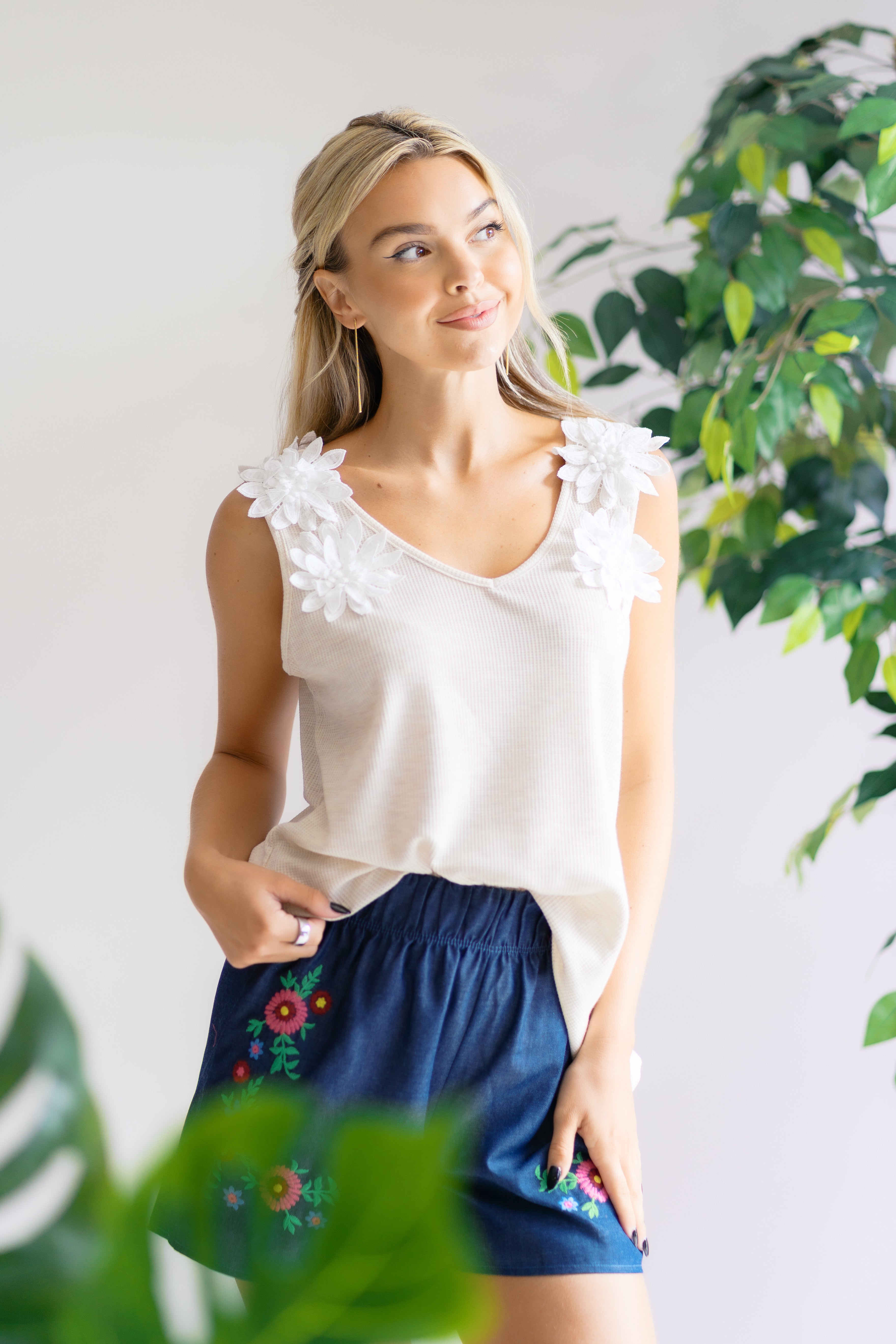 Woman wearing a white blouse with floral details and blue shorts with embroidery, standing against a light background with greenery.