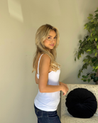 Woman in a white tank top and jeans standing indoors with a plant in the background