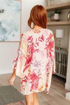 Woman wearing a floral kimono-style garment in a room with a painting and lamp in the background.