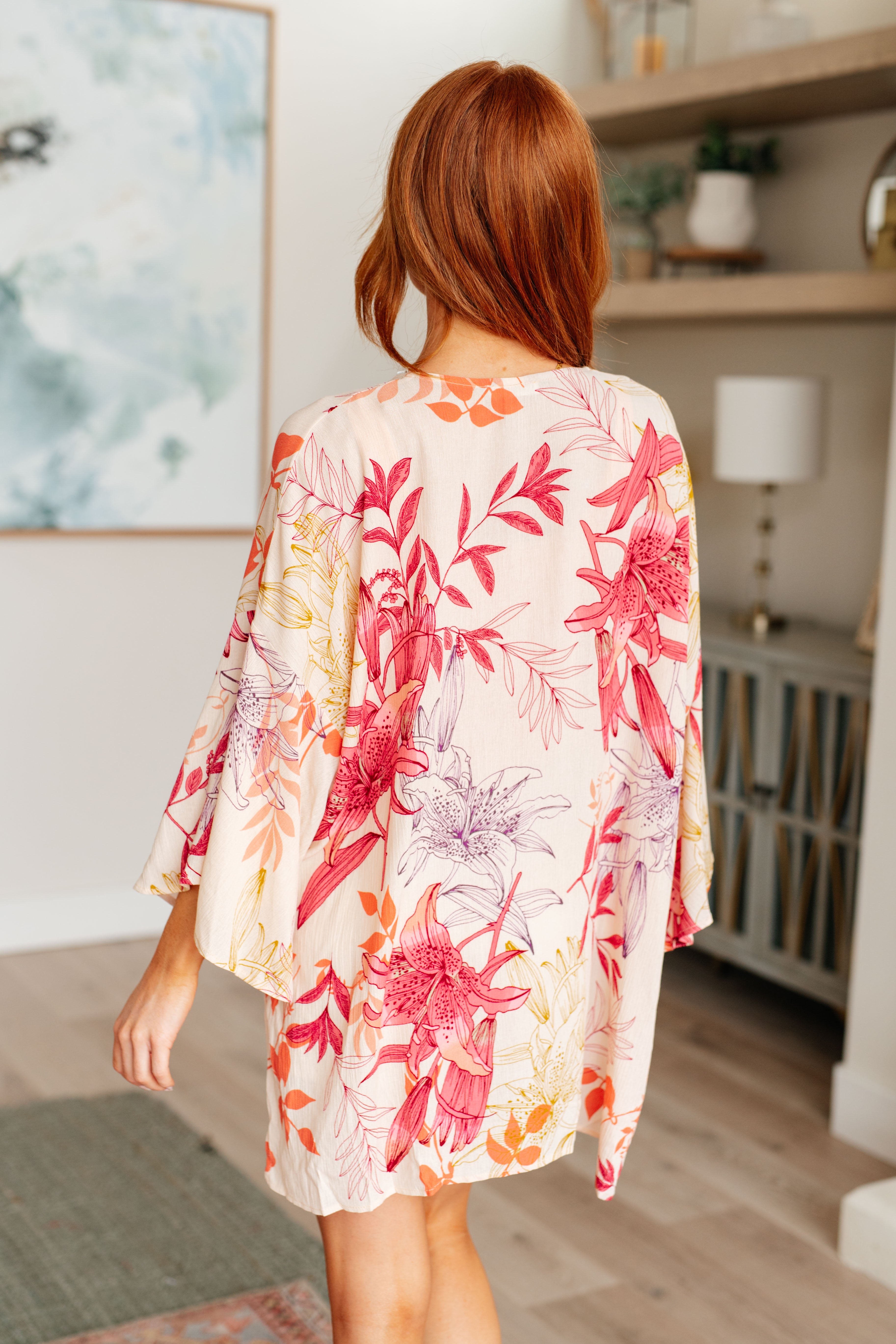 Woman wearing a floral kimono-style garment in a room with a painting and lamp in the background.
