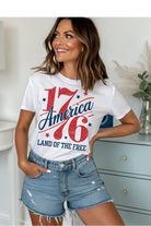 Woman wearing a white t-shirt with '1776 America Land of the Free' design in a casual setting.