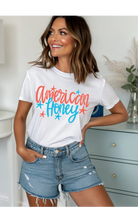Woman wearing a white t-shirt with 'American Honey' text, standing in a room.